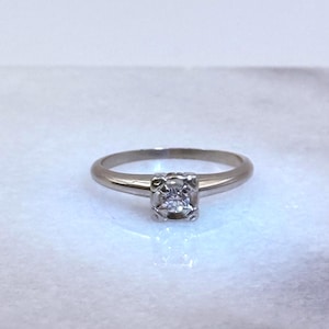 May include: A silver engagement ring with a square-shaped diamond set in a four-prong setting. The ring has a thin band and is displayed on a white surface. The ring is a classic style.