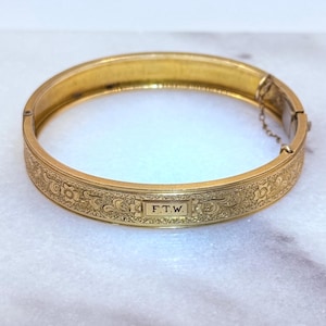 May include: A gold-colored hinged bangle bracelet. The bracelet features an engraved floral pattern and the initials "F.T.W." on a rectangular plaque. The bracelet is approximately 0.75 inches wide.