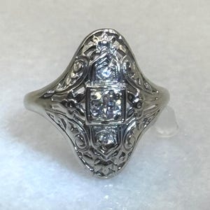 May include: A silver-toned ring with an ornate filigree design. The ring features a central square setting with a clear gemstone, flanked by two smaller gemstones. The ring's band has a delicate, flowing shape.