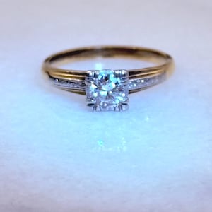 May include: A gold and silver engagement ring with a large, square-cut diamond center stone. The band is two-toned, with a gold exterior and a silver interior. Small accent stones are set along the silver band, adding sparkle to the ring.