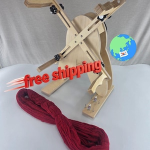 May include: A light-colored wooden yarn swift with adjustable arms and black knobs, featuring a circular base and a metal spindle mechanism. A large, twisted hank of deep red yarn lies in the foreground. A globe icon with a South Korean flag is visible.