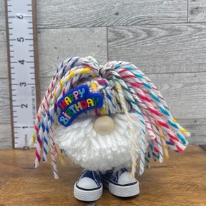 May include: A gnome figurine with a white yarn beard, wearing a hat made of multi-colored yarn strands. A blue plastic emblem with the text "HAPPY BIRTHDAY" is attached to the hat. The gnome wears blue and white canvas sneakers.