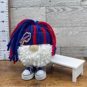 May include: A gnome figurine wearing a blue and red striped yarn hat with a Buffalo Bills logo pin, a white yarn beard, and blue and white sneakers. A ruler visible on the left shows the gnome stands approximately 4.5 inches tall.