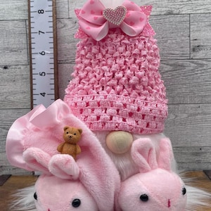 May include: A pink Easter gnome with bunny ears and a pink crocheted hat. The gnome has a white beard, a wooden nose, and a pink bow with a heart-shaped rhinestone center. A small teddy bear is attached to the bunny ears.