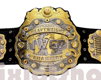 Pode incluir: Um cinturão de campeonato dourado e prateado com uma placa central grande que diz "IWGP Heavyweight Champion". O cinturão tem uma alça de couro preto com várias placas menores que mostram os nomes dos campeões anteriores.