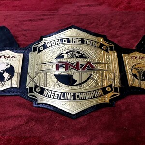 Könnte beinhalten: Ein goldener und schwarzer Wrestling-Meisterschaftsgürtel mit den Worten "World Tag Team Wrestling Champion" und "TNA" darauf. Der Gürtel hat ein Globus-Design und schwarze Lederriemen.