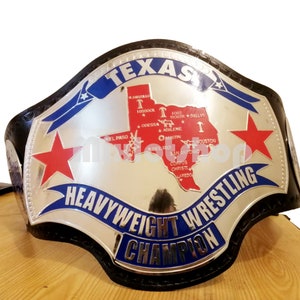Könnte beinhalten: Ein silberner Meisterschaftsgürtel mit blauem und rotem Design. Der Gürtel zeigt eine Karte von Texas mit Städtenamen und einem großen Stern auf jeder Seite. Der Text "TEXAS HEAVYWEIGHT WRESTLING CHAMPION" ist auf dem Gürtel geschrieben.
