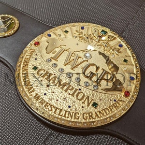 Könnte beinhalten: Ein goldener Meisterschaftsgürtel mit einer großen, verzierten Mittelplatte, die eine Weltkarte, den Text "IWGP" und die Worte "International Wrestling Grand Prix Champion" zeigt.