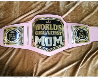 May include: A pink championship belt with gold accents and rhinestones. The center plate reads "World's Greatest Mom" with a crown above the text. The side plates read "Best Mom Ever" with a crown above the text.