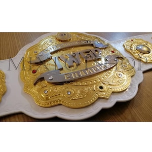 Könnte beinhalten: Ein weißer Wrestling-Championschaftsgürtel mit goldenen und silbernen Akzenten. Der Gürtel trägt die Worte "IWE" und "Champion" auf der Mittelplatte.