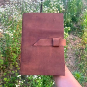 Può includere: Un diario in pelle marrone con chiusura a cinturino. Il diario è rettangolare e sembra fatto a mano. La pelle ha un aspetto leggermente invecchiato e il cinturino è fissato con un anello.