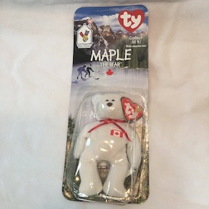 May include: White Ty Beanie Baby plush toy named "Maple the Bear" with a red ribbon and a Canadian flag patch. The packaging features a mountain scene and the text "Collect All 4! While supplies last."