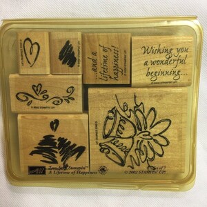 May include: A set of seven wooden stamps in a clear plastic case. The stamps feature various designs, including hearts, swirls, bells, and the phrase "...and a lifetime of happiness!" The stamps are part of the "Two-Step Stampin' A Lifetime of Happiness" collection.