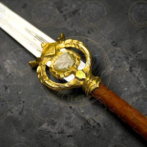 Puede incluir: Primer plano de una espada de fantasía con una empuñadura dorada y una piedra transparente en forma de gema en el centro. La hoja es plateada y el mango está envuelto en cuero marrón. La espada está sobre un fondo oscuro y texturizado.