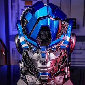 Peut inclure: Buste métallique détaillé de robot, principalement bleu et argenté. Le design présente des éléments mécaniques complexes, des yeux bleus brillants et un emblème central. L'esthétique suggère un thème de science-fiction ou de figurine d'action.