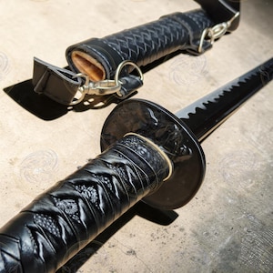 May include: A black katana sword with a textured handle and a black scabbard. The sword blade is black with a silver edge. The handle is wrapped in a woven pattern. The scabbard has a leather strap and a gold-colored clasp.