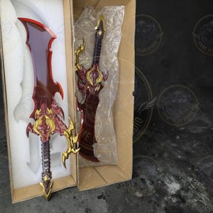 May include: Two ornate fantasy swords in cardboard boxes. The swords have red blades and gold and purple accents. One sword is partially visible in its box, while the other is fully displayed. The swords are likely props or collectibles.