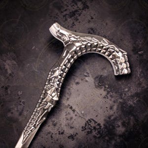 May include: Silver-colored walking cane with intricate, geometric patterns and a curved handle. The handle features a textured, diamond-like design. The cane's shaft has detailed, sculpted embellishments, creating a decorative and functional accessory.