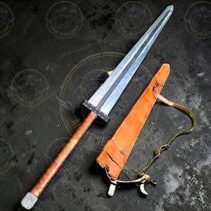 May include: A large, silver and black sword with a brown leather-wrapped handle and a matching orange sheath. The blade has a central dark stripe. The sheath has a leather strap and a metal clasp.