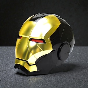 Może przedstawiać: Złoty i czarny hełm Iron Mana z czerwonymi szczelinami na oczy. Hełm jest repliką kultowego nakrycia głowy superbohatera, z metalicznym złotym wykończeniem na twarzy i czarnym wykończeniem z tyłu i po bokach.