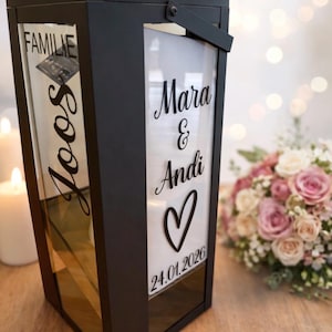 Könnte beinhalten: Eine schwarze Metalllaterne mit Glasscheiben und schwarzer Vinylbeschriftung. Eine Seite zeigt den Text "FAMILIE Joos", die andere Seite "Mara & Andi" mit einem Herzsymbol und dem Datum "24.01.2026" in einer geschwungenen Schreibschrift.