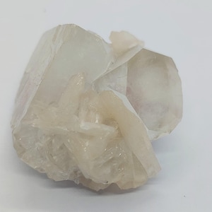 Puede incluir: Un grupo de cristales minerales translúcidos de color blanco roto con formaciones geométricas angulares y afiladas y una textura suave y vítrea. El espécimen muestra una combinación de estructuras cristalinas más grandes y bloques junto a segmentos más pequeños y dentados.