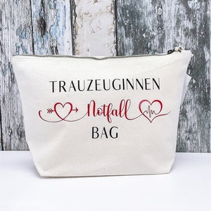 May include: White canvas makeup bag with a zipper closure. The bag has black text that reads "Trauzeuginnen Notfall Bag" with a red heart and heartbeat symbol on either side of the text.