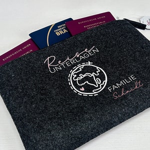 Könnte beinhalten: Eine dunkelgraue Filz-Reisedokumententasche mit Reißverschluss, die mehrere Pässe enthält. Der Halter hat weißen und rosa Text mit der Aufschrift "Reise Unterlagen Familie Schmidt" mit einer Weltkarten-Grafik. Ein kleines Etikett mit floralen Illustrationen ist angebracht.