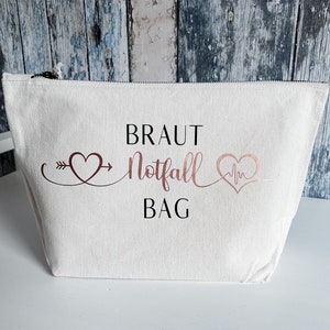 May include: White canvas zippered pouch with the text "Braut Notfall Bag" in rose gold lettering. The text is surrounded by two hearts, one with an arrow and the other with a heartbeat symbol.