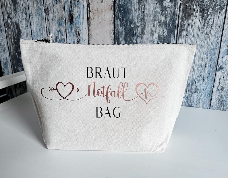 May include: White canvas zippered pouch with the text "Braut Notfall Bag" in rose gold lettering. The text is surrounded by two hearts, one with an arrow and the other with a heartbeat symbol.