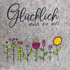 Puede incluir: Bolsa de tela gris con el texto "Gl&uuml;cklich steht dir gut!" en letras negras. La bolsa presenta una fila de flores coloridas con acentos amarillos, rosas, rojos y morados.