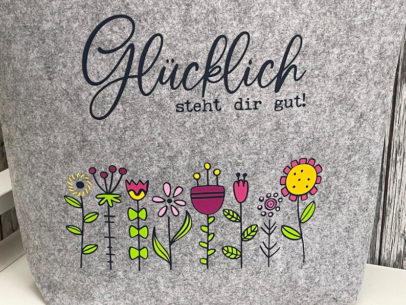 Puede incluir: Bolsa de tela gris con el texto "Gl&uuml;cklich steht dir gut!" en letras negras. La bolsa presenta una fila de flores coloridas con acentos amarillos, rosas, rojos y morados.