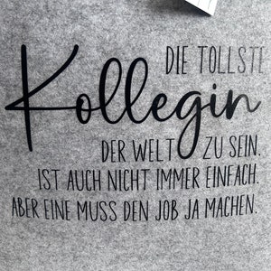 May include: Gray t-shirt with black text that reads "DIE TOLLSTE Kollegin DER WELT ZU SEIN. IST AUCH NICHT IMMER EINFACH. ABER EINE MUSS DEN JOB JA MACHEN."