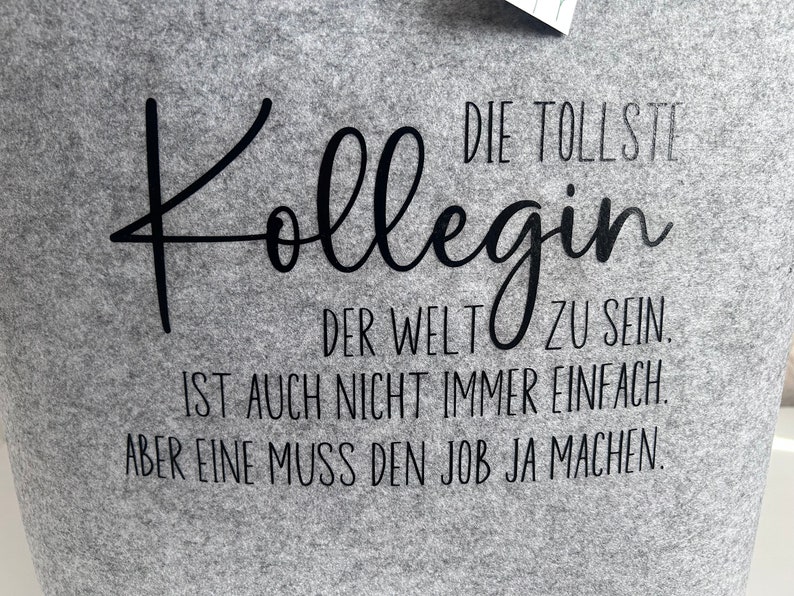 May include: Gray t-shirt with black text that reads "DIE TOLLSTE Kollegin DER WELT ZU SEIN. IST AUCH NICHT IMMER EINFACH. ABER EINE MUSS DEN JOB JA MACHEN."
