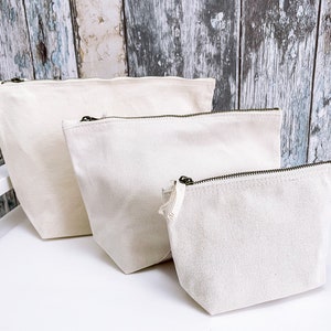 May include: Three off-white canvas zippered pouches of different sizes. The pouches are lined up from largest to smallest on a white surface.