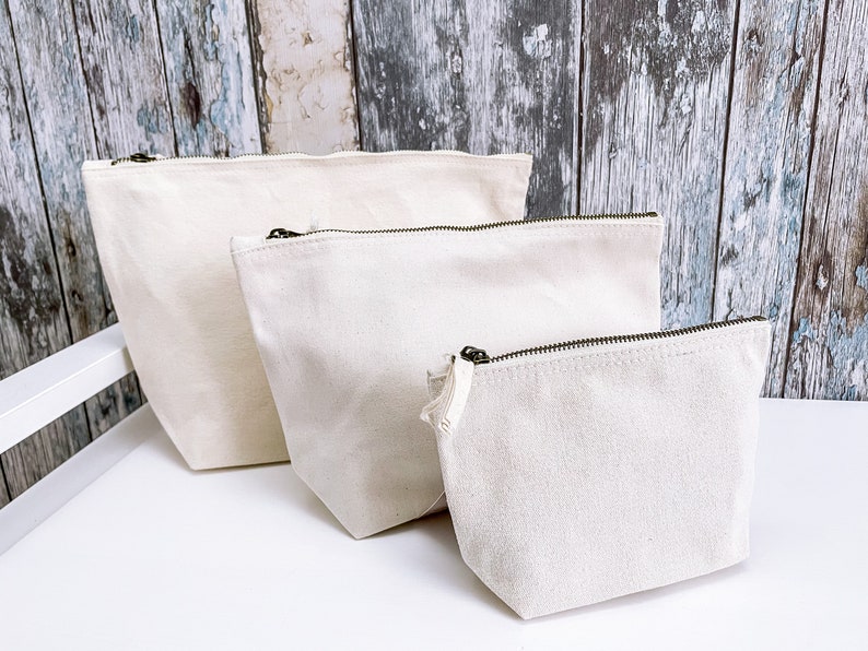 May include: Three off-white canvas zippered pouches of different sizes. The pouches are lined up from largest to smallest on a white surface.