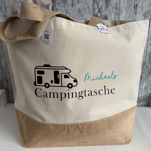 Könnte beinhalten: Beige Canvas-Tragetasche mit braunem Juteboden und Griffen. Die Tasche hat eine schwarze Grafik eines Wohnmobils und die Wörter "Campingtasche" und "Michaels" in Türkis.