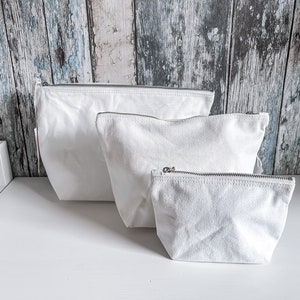 May include: Three white canvas zippered pouches of different sizes. The pouches are lined up on a white surface.