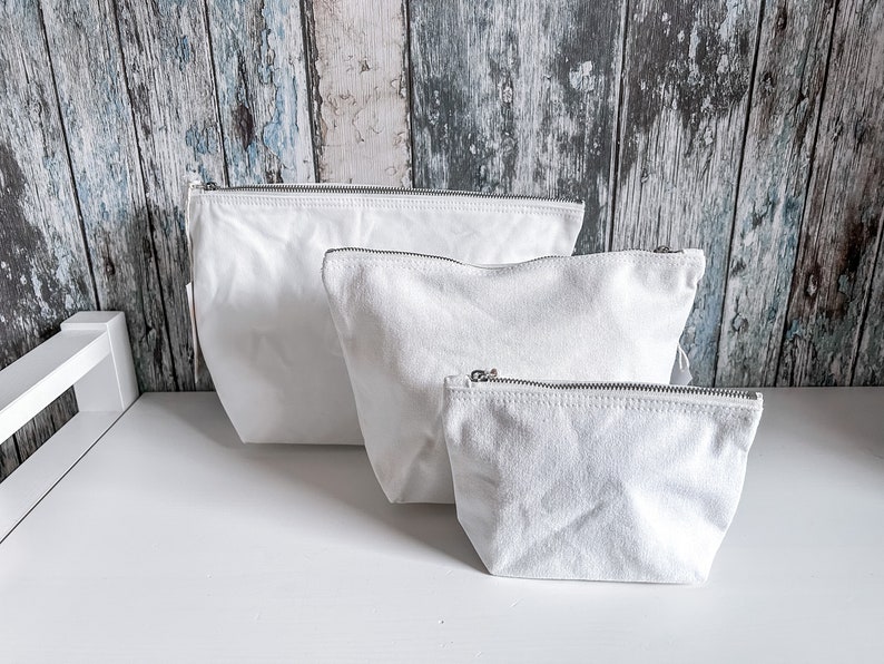 May include: Three white canvas zippered pouches of different sizes. The pouches are lined up on a white surface.