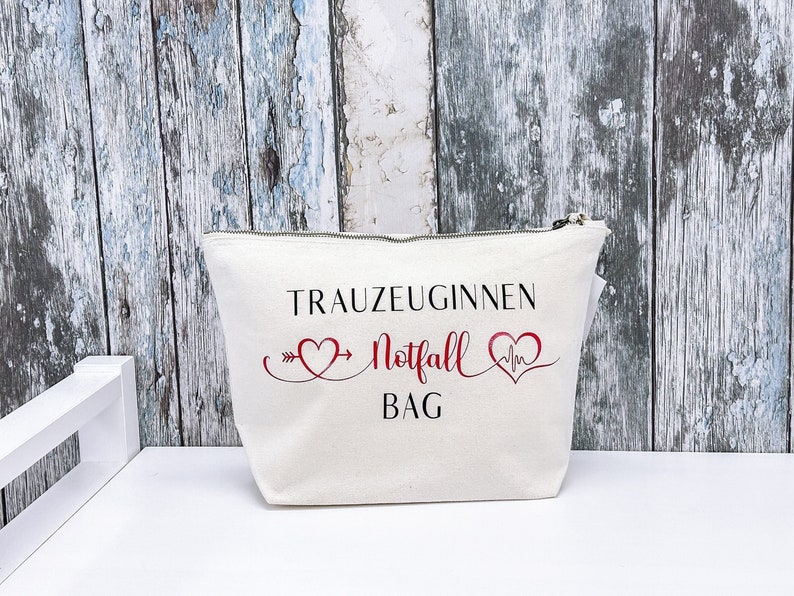 May include: White canvas makeup bag with a zipper closure. The bag has black text that reads "Trauzeuginnen Notfall Bag" with a red heart and heartbeat symbol on either side of the text.