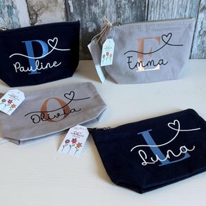 May include: Four personalized canvas zipper pouches in navy and gray. Each pouch features a name and initial in various colors, with a heart design. Small tags with floral designs are attached to some of the pouches.