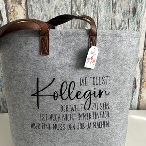 May include: Gray felt tote bag with brown leather handles. The bag has black text that reads "DIE TOLLSTE KOLLEGIN DER WELT ZU SEIN IST AUCH NICHT IMMER EINFACH. ABER EINE MUSS DEN JOB JA MACHEN."