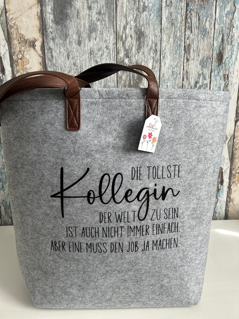 May include: Gray felt tote bag with brown leather handles. The bag has black text that reads "DIE TOLLSTE KOLLEGIN DER WELT ZU SEIN IST AUCH NICHT IMMER EINFACH. ABER EINE MUSS DEN JOB JA MACHEN."