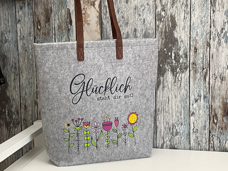 Puede incluir: Bolsa de tela gris con asas de cuero marr&oacute;n. La bolsa tiene un gr&aacute;fico en blanco y negro de flores y el texto "Gl&uuml;cklich steht dir gut!"