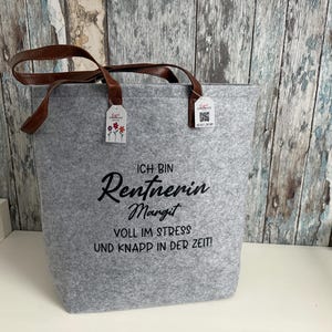 Könnte beinhalten: Graue Filz-Tragetasche mit braunen Ledergriffen. Die Tasche trägt den deutschen Text "ICH BIN Rentnerin Margit VOLL IM STRESS UND KNAPP IN DER ZEIT!" und kleine Anhänger mit floralen Designs.