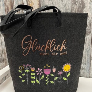 Puede incluir: Una bolsa de tela gris con asas negras. La bolsa tiene una inscripci&oacute;n en oro rosa que dice "Gl&uuml;cklich steht dir gut!" y una fila de flores de colores en la parte inferior.