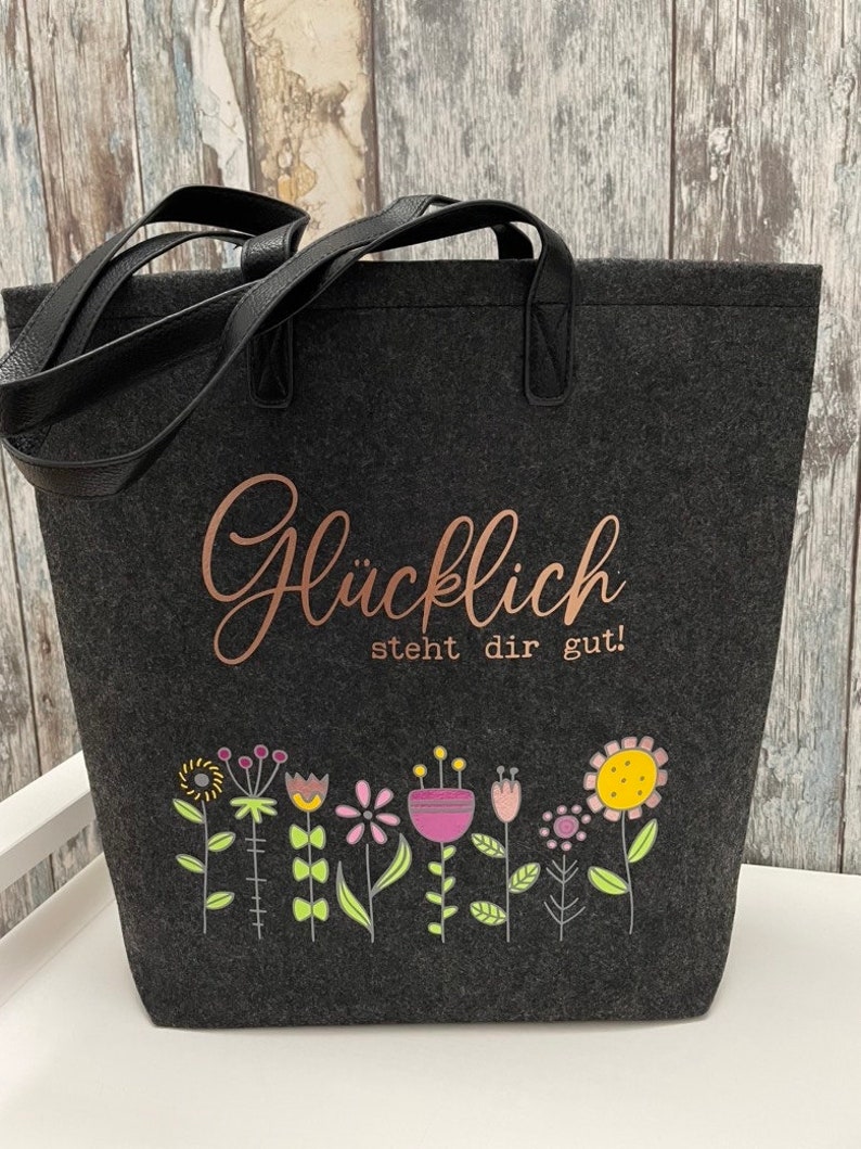 Puede incluir: Una bolsa de tela gris con asas negras. La bolsa tiene una inscripci&oacute;n en oro rosa que dice "Gl&uuml;cklich steht dir gut!" y una fila de flores de colores en la parte inferior.