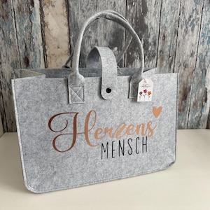 Könnte beinhalten: Eine graue Filztasche mit einer roségoldenen Aufschrift "Herzens Mensch".