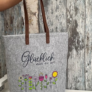 Puede incluir: Bolsa de tela gris con asas de cuero marr&oacute;n. La bolsa tiene un gr&aacute;fico en blanco y negro de flores y el texto "Gl&uuml;cklich steht dir gut!"
