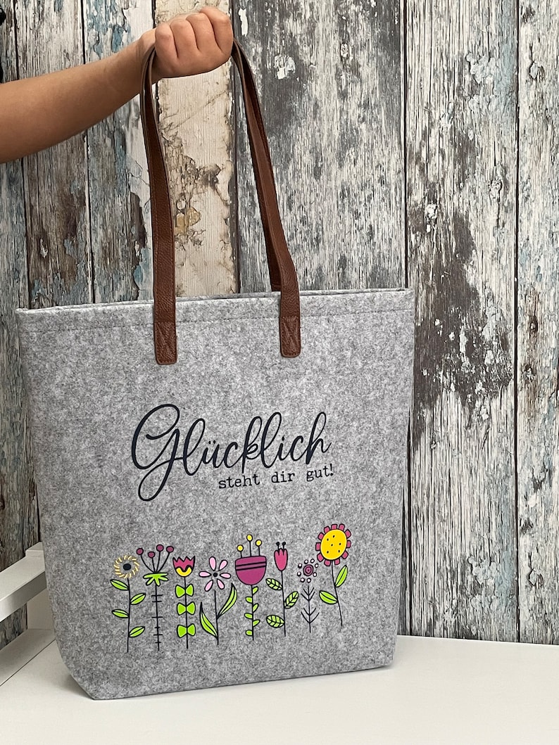 Puede incluir: Bolsa de tela gris con asas de cuero marr&oacute;n. La bolsa tiene un gr&aacute;fico en blanco y negro de flores y el texto "Gl&uuml;cklich steht dir gut!"